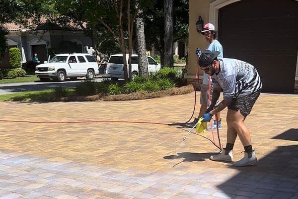 Two men clean a brick driveway with a power washer. One man sprays the bricks while the other holds the hose. White SUV parked on the lawn of a home in the background.
