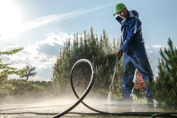 Man in blue protective suit power washing a patio outdoors. Water spray, green hat, and coiled hose are visible. Maintaining your Home with pressure washing.